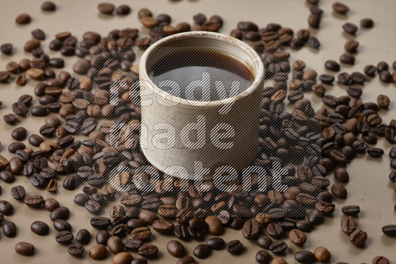 A beige pottery cup of coffee surrounded by roasted coffee beans on beige background