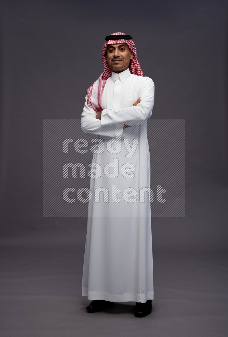 Saudi man wearing thob and shomag standing with crossed arms on gray background