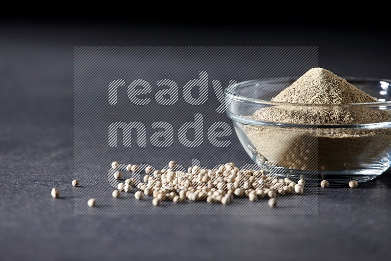 A glass bowl full of white pepper powder with white pepper beads on black flooring