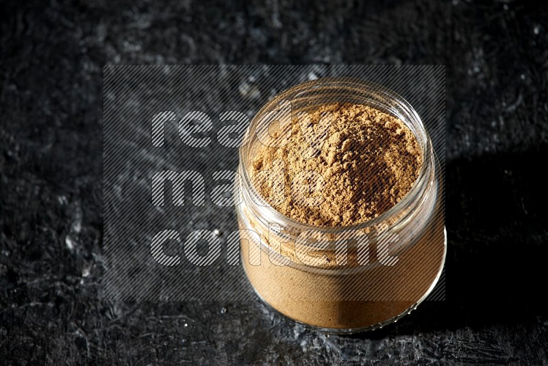 A glass jar full of allspice powder on a textured black flooring