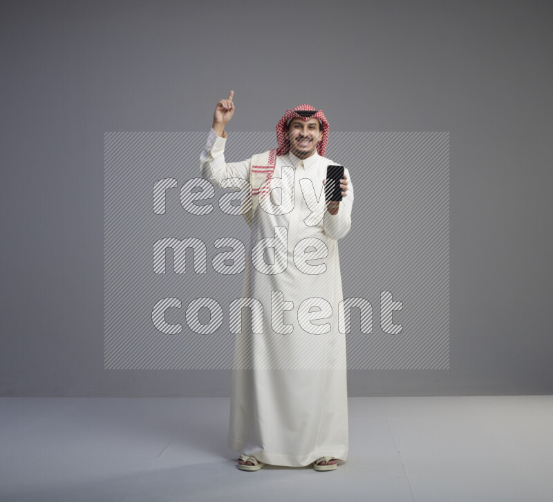 A Saudi man standing wearing thob and red shomag showing phone to camera on gray background