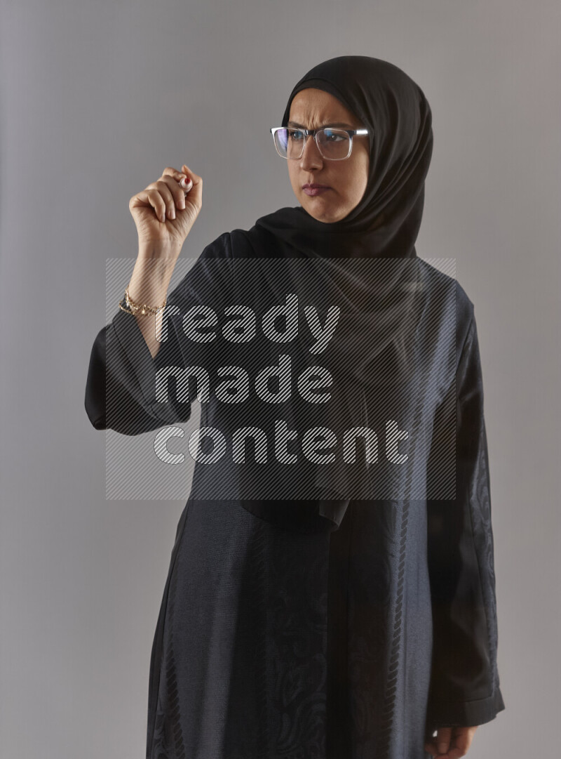 Woman in black abaya, hijab and glasses holding a marker pen to write on a transparent board with different reactions and expressions on grey background