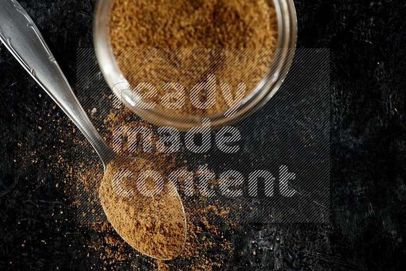 A glass jar and a metal spoon full of cumin powder on a textured black flooring