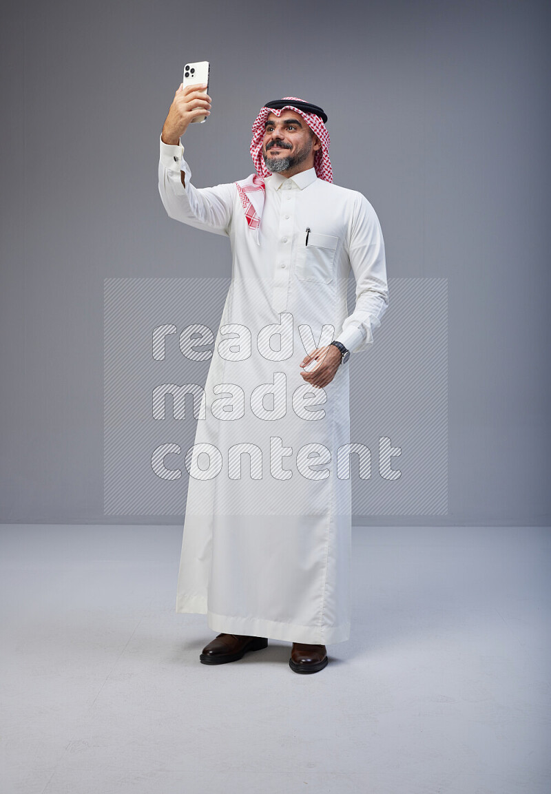 Saudi man Wearing Thob and red Shomag standing taking selfie on Gray background