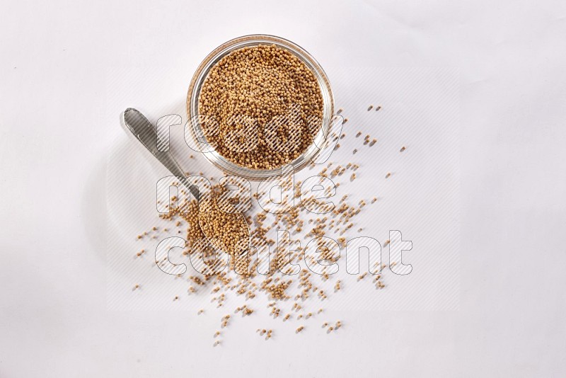 A glass bowl and metal spoon full of mustard seeds on a white flooring
