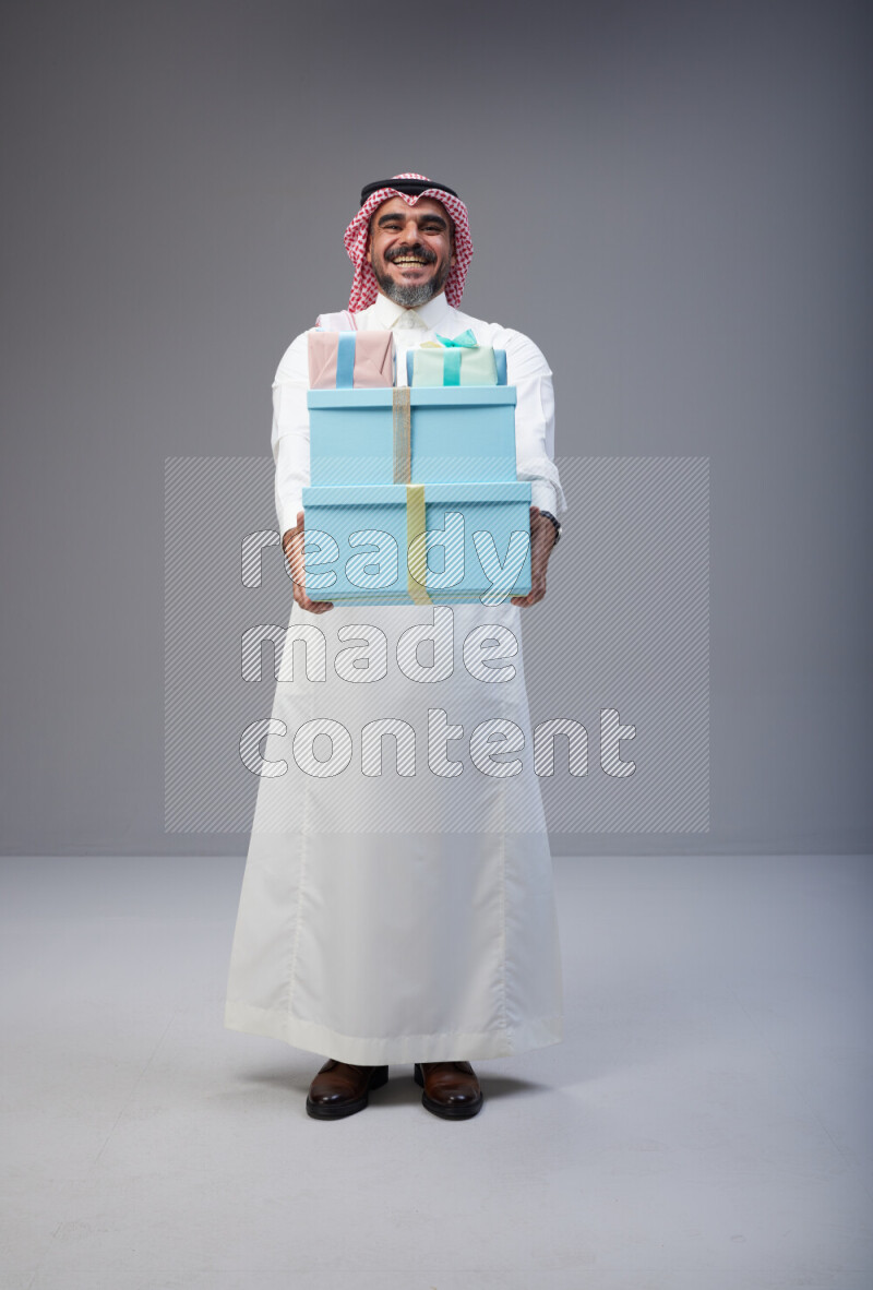 Saudi man Wearing Thob and red Shomag standing holding gift box on Gray background
