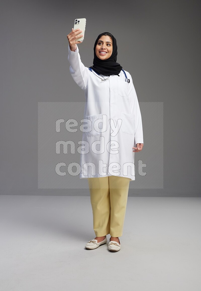 Saudi woman wearing lab coat with stethoscope standing taking selfie on Gray background