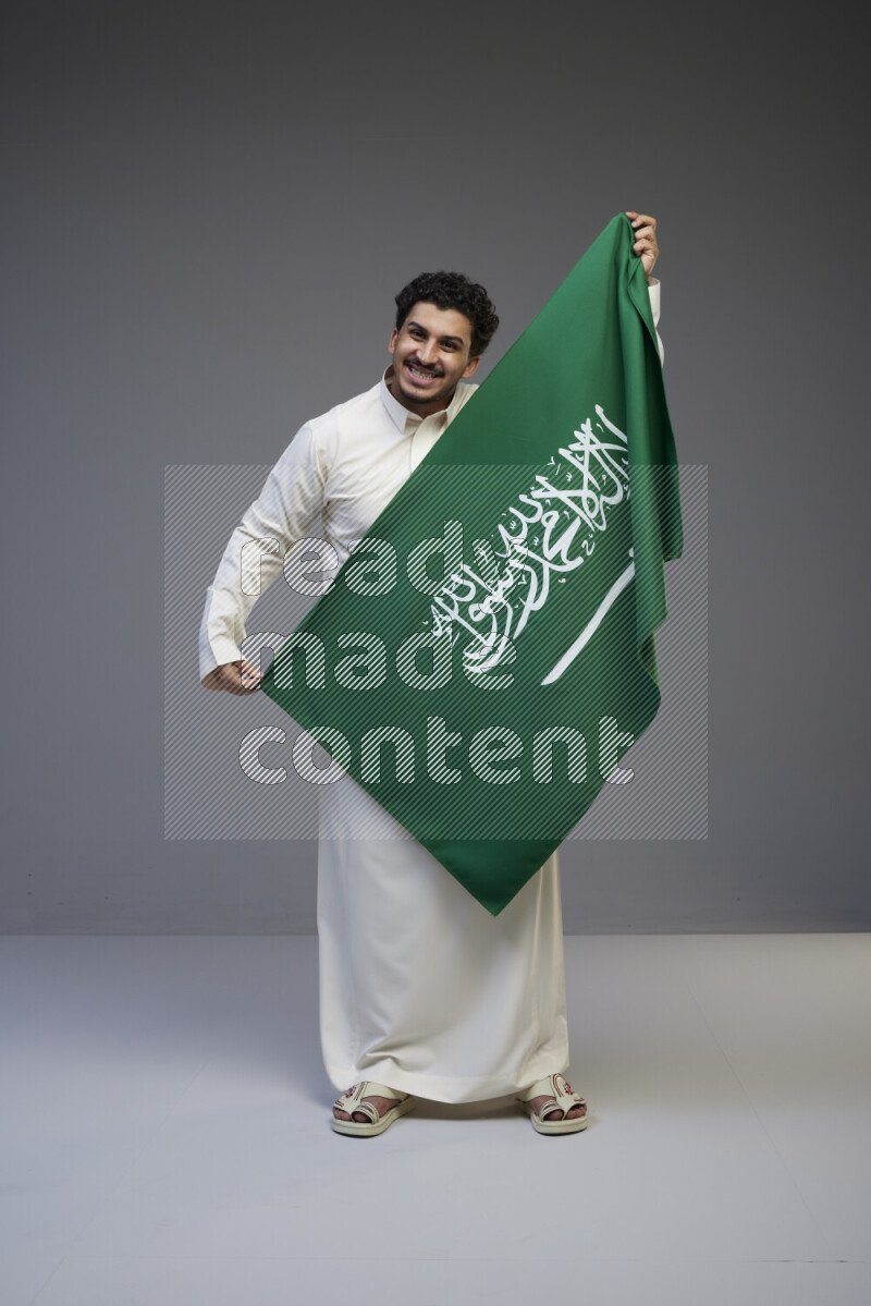A Saudi man standing wearing thob holding big Saudi flag on gray background