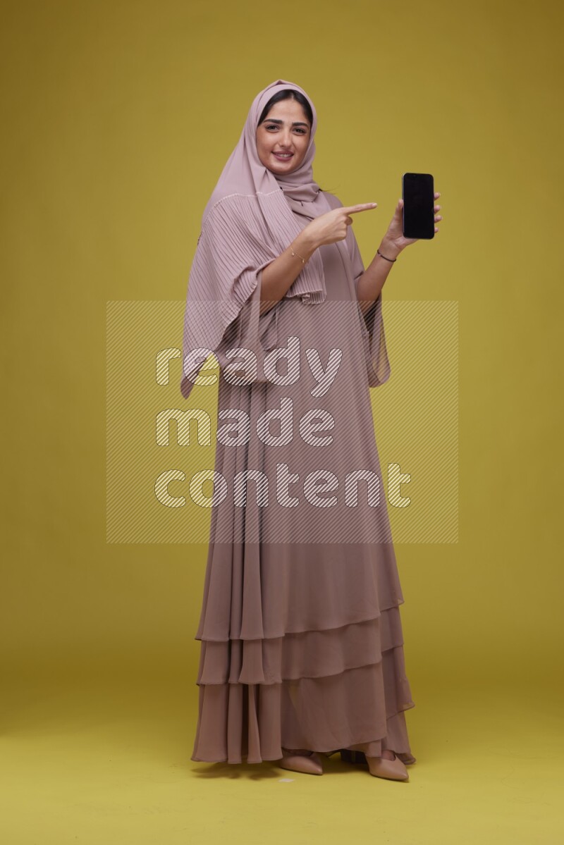 A woman Showing her Phone screen on a Yellow Background wearing Brown Abaya with Hijab