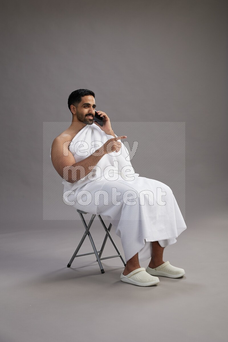 A man wearing Ehram sitting on chair talking on the phone on gray background