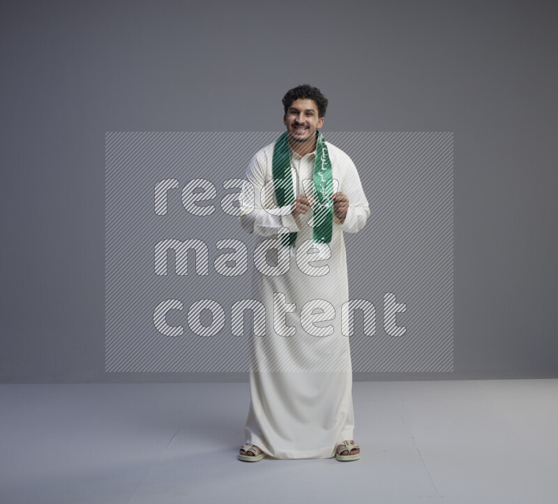 A Saudi man standing wearing thob and Saudi flag scarf on gray background
