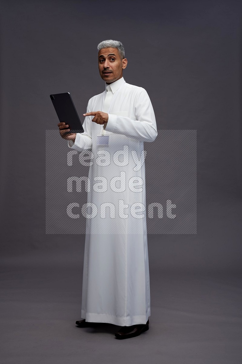Saudi man wearing thob with neck strap employee badge standing working on tablet on gray background
