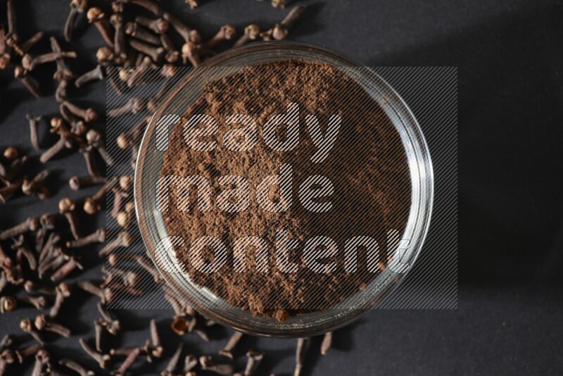 A glass bowl full of cloves powder with cloves grains spread on black flooring