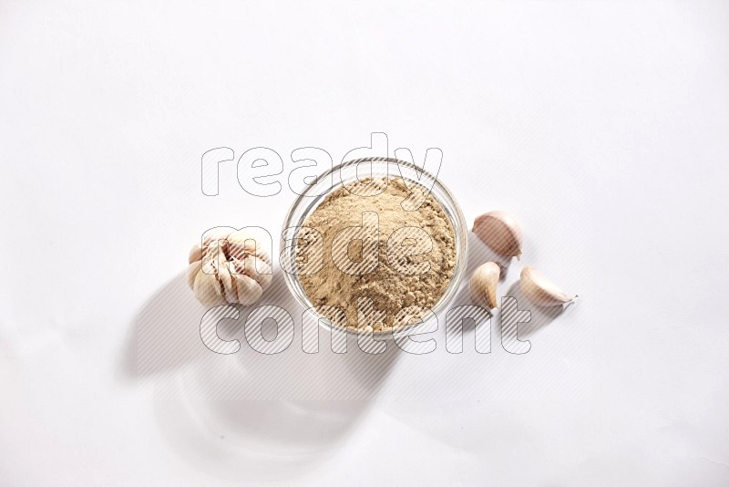 A glass bowl full of garlic powder and beside it garlic bulb and cloves on a white flooring in different angles