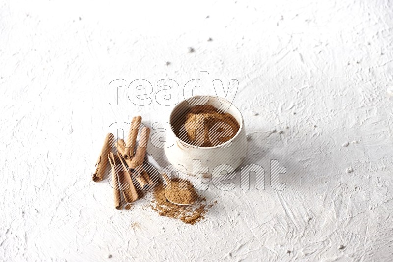 Ceramic beige bowl full of cinnamon powder and a metal spoon with cinnamon sticks next of it on a textured white background