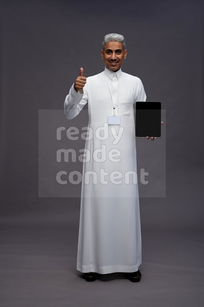 Saudi man wearing thob with neck strap employee badge standing showing tablet to camera on gray background