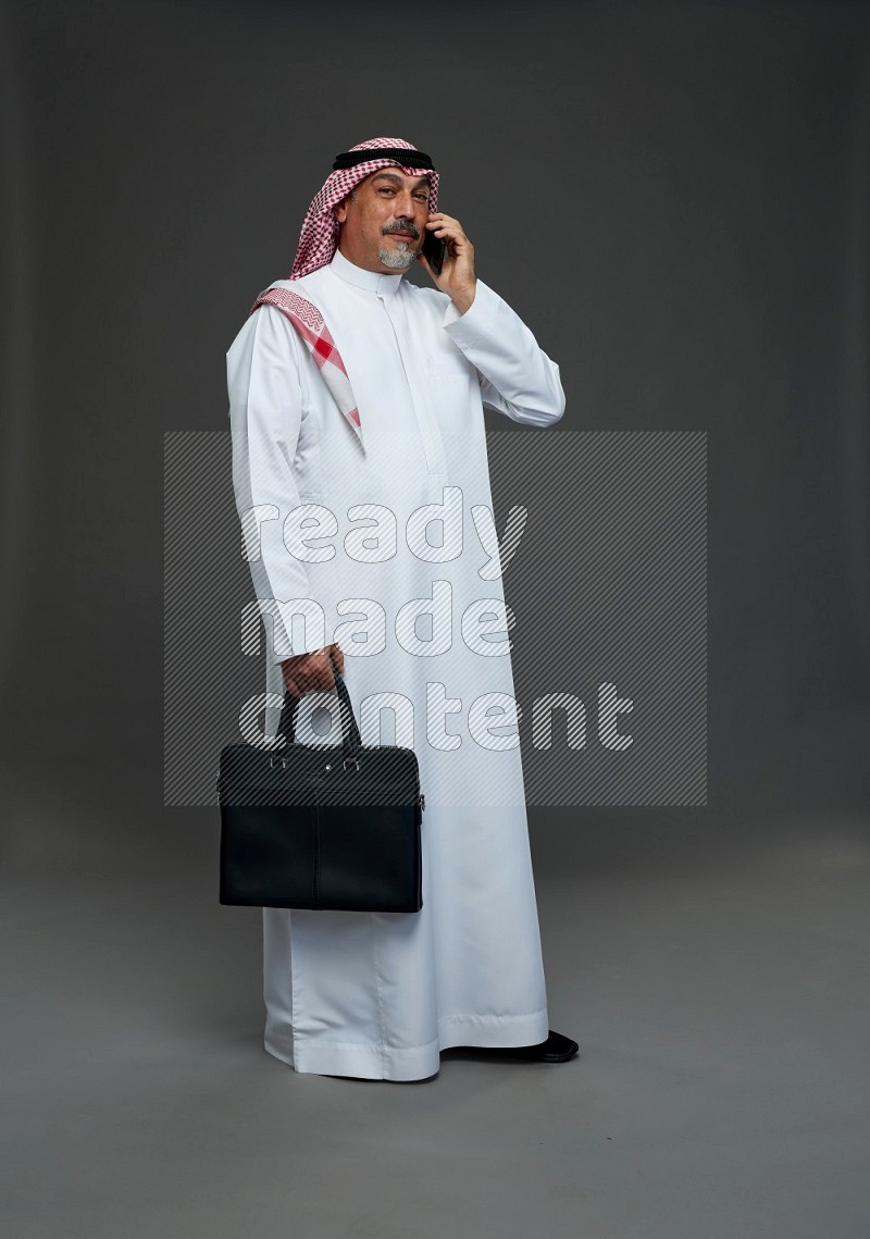 Saudi man with shomag Standing holding bag talking on phone on gray background