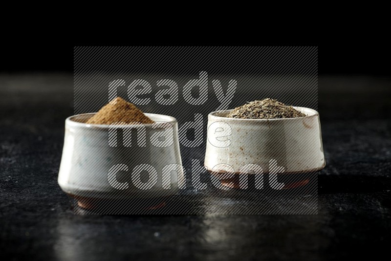 2 beige bowls, one full of cumin powder and the other full of seeds on a textured black flooring