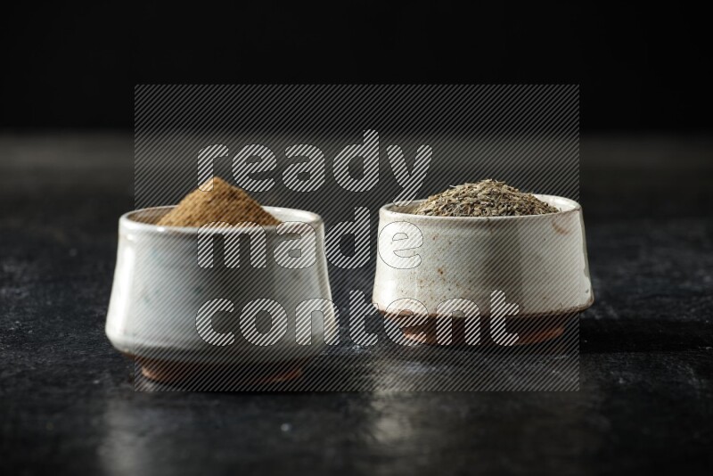 2 beige bowls, one full of cumin powder and the other full of seeds on a textured black flooring