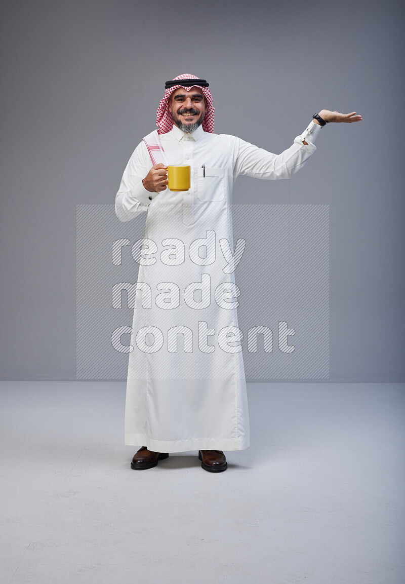 Saudi man Wearing Thob and red Shomag standing holding mug on Gray background