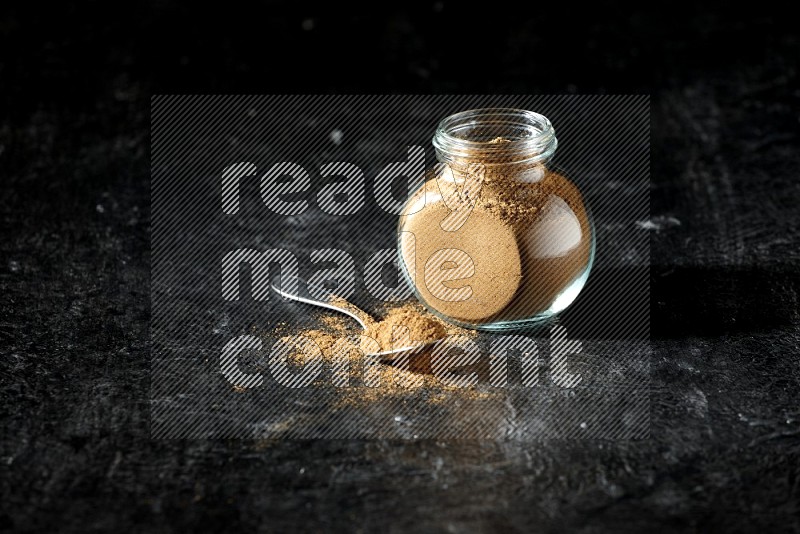 A glass spice jar and metal spoon full of allspice powder on a textured black flooring