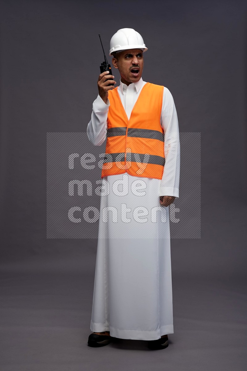 Saudi man wearing thob with engineer vest standing holding walkie-talkie on gray background