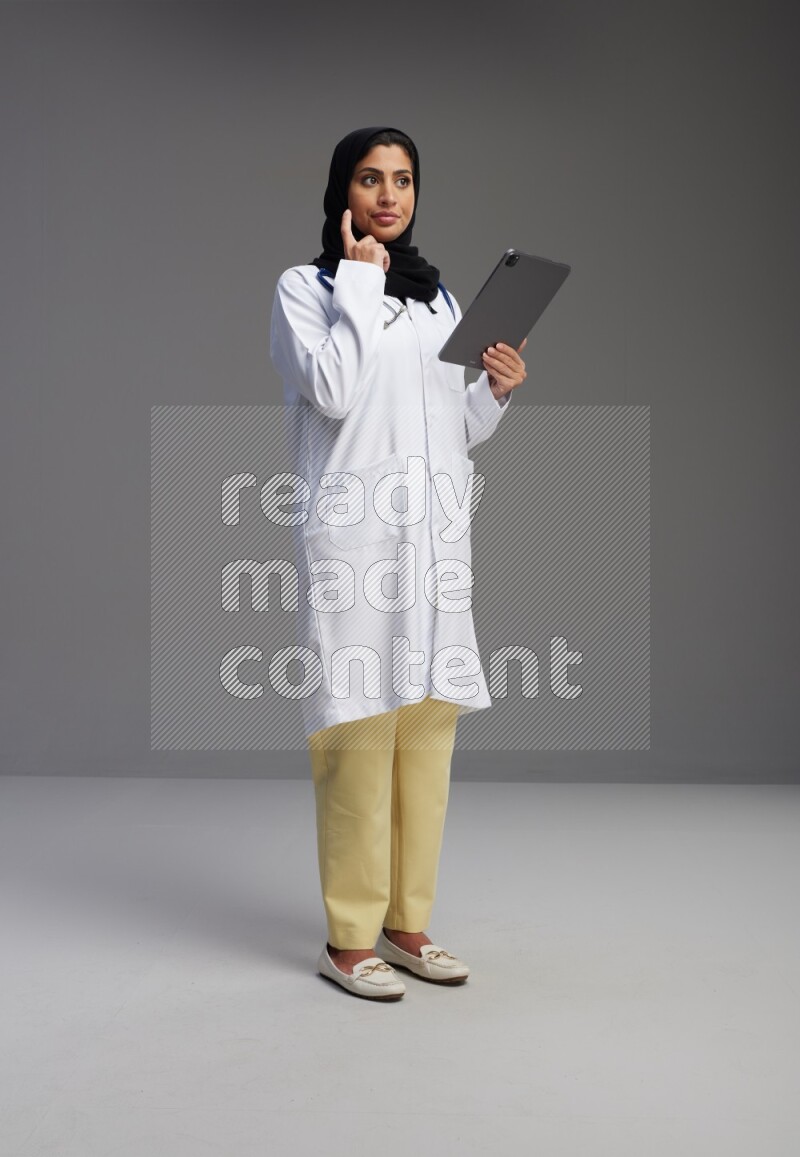 Saudi woman wearing lab coat with stethoscope standing working on tablet on Gray background