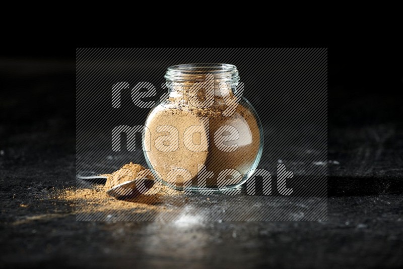 A glass spice jar and metal spoon full of allspice powder on a textured black flooring