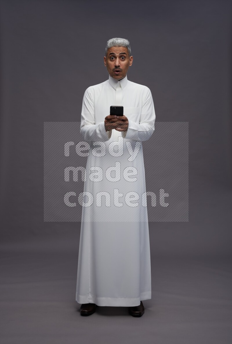 Saudi man wearing thob standing texting on phone on gray background