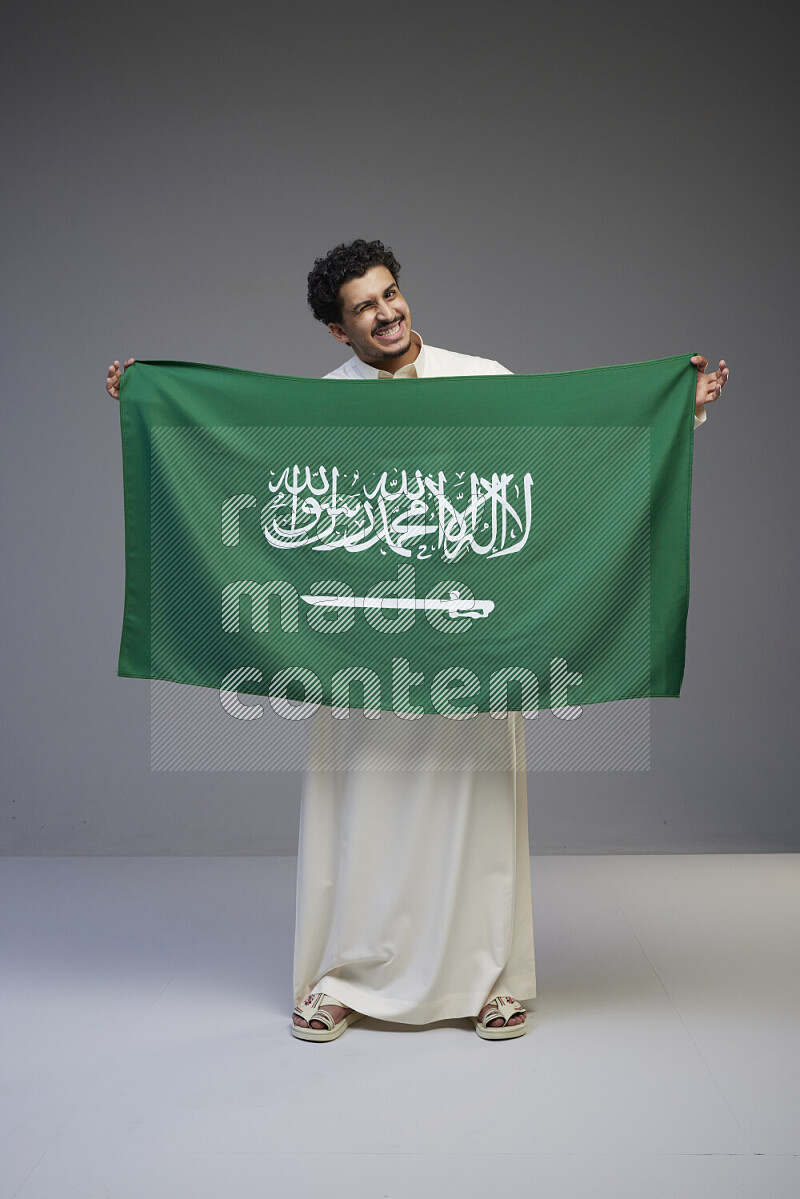 A Saudi man standing wearing thob holding big Saudi flag on gray background