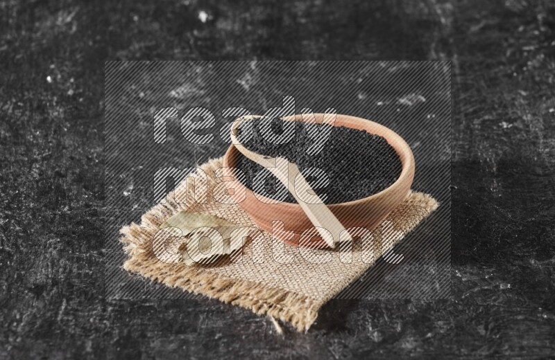 وعاء خشبي مع ملعقة خشبية ممتلئان ببذور حبة البركة علي قطعة من القماش علي خلفية سوداء