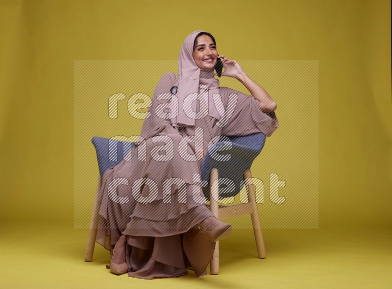 A woman having a call on a blue Chair on a Yellow Background wearing Brown Abaya with Hijab