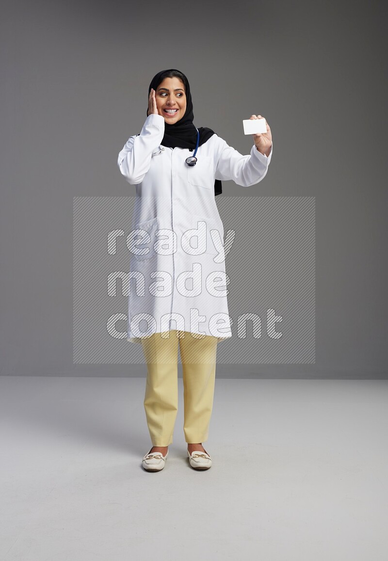 Saudi woman wearing lab coat with stethoscope standing holding ATM card on Gray background