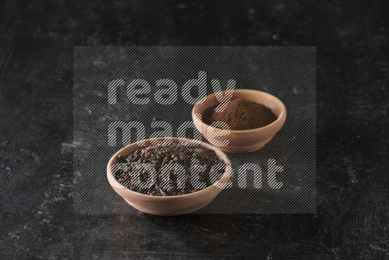 2 wooden bowls full of cloves powder and whole cloves on a textured black flooring