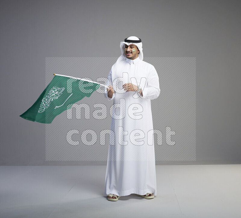 A Saudi man standing wearing thob and white shomag with face painting raising big Saudi flag on gray background