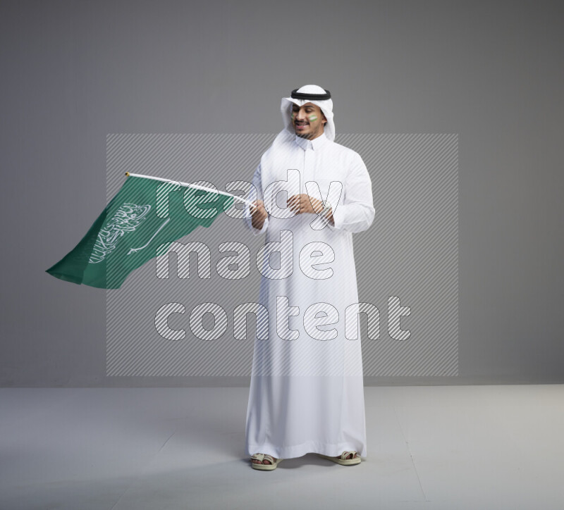 A Saudi man standing wearing thob and white shomag with face painting raising big Saudi flag on gray background