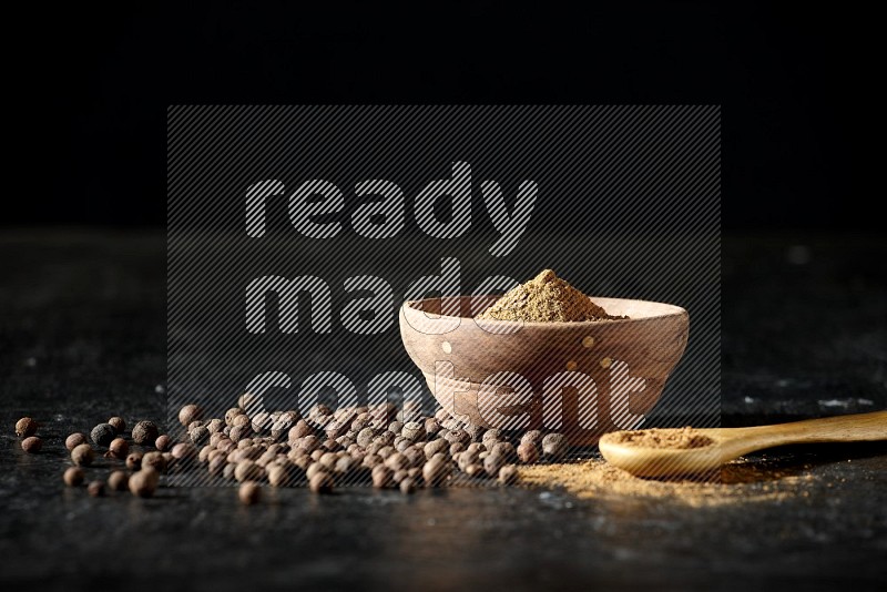 A wooden bowl and spoon full of allspice powder and whole balls spreaded on a black flooring