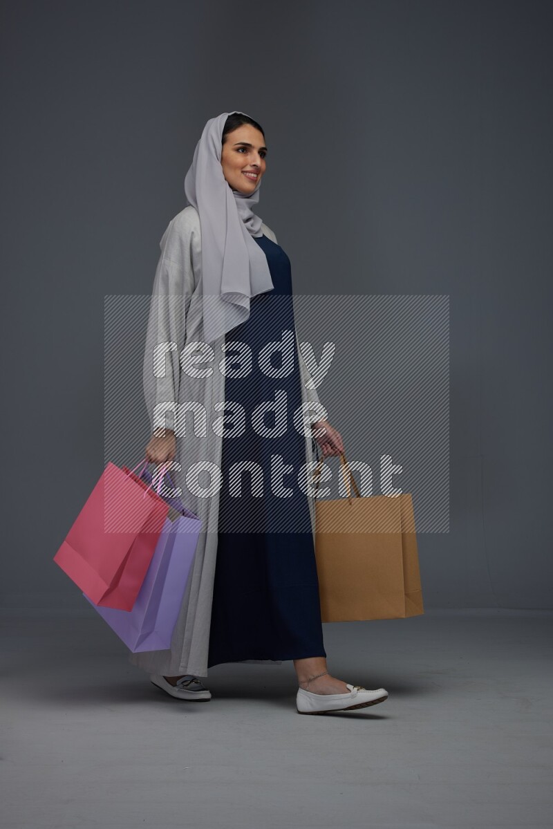 A Saudi woman wearing a light gray Abaya and head scarf standing and holding shopping bags on a grey background