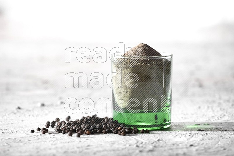 A glass cup full of black pepper powder and black pepper spread on a textured white flooring