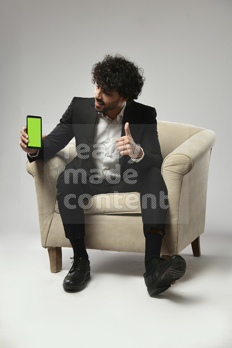 A man wearing formal sitting on a chair showing a phone screen on white background
