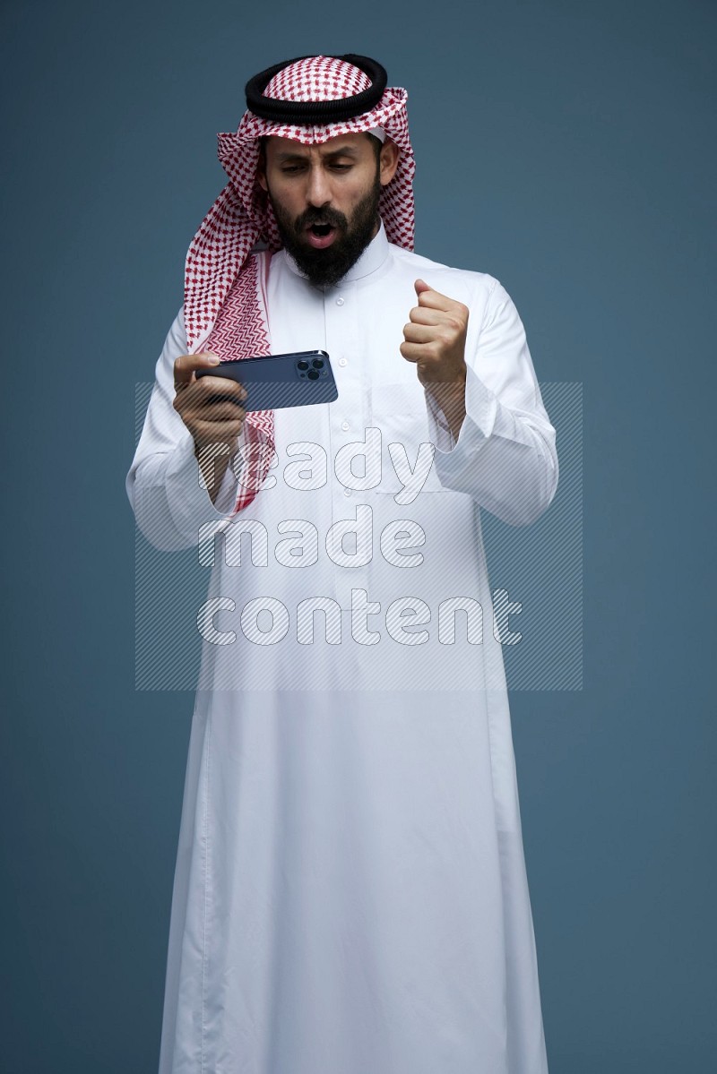 A man Playing a Game on his phone in a blue background wearing Saudi Thob with Shomag