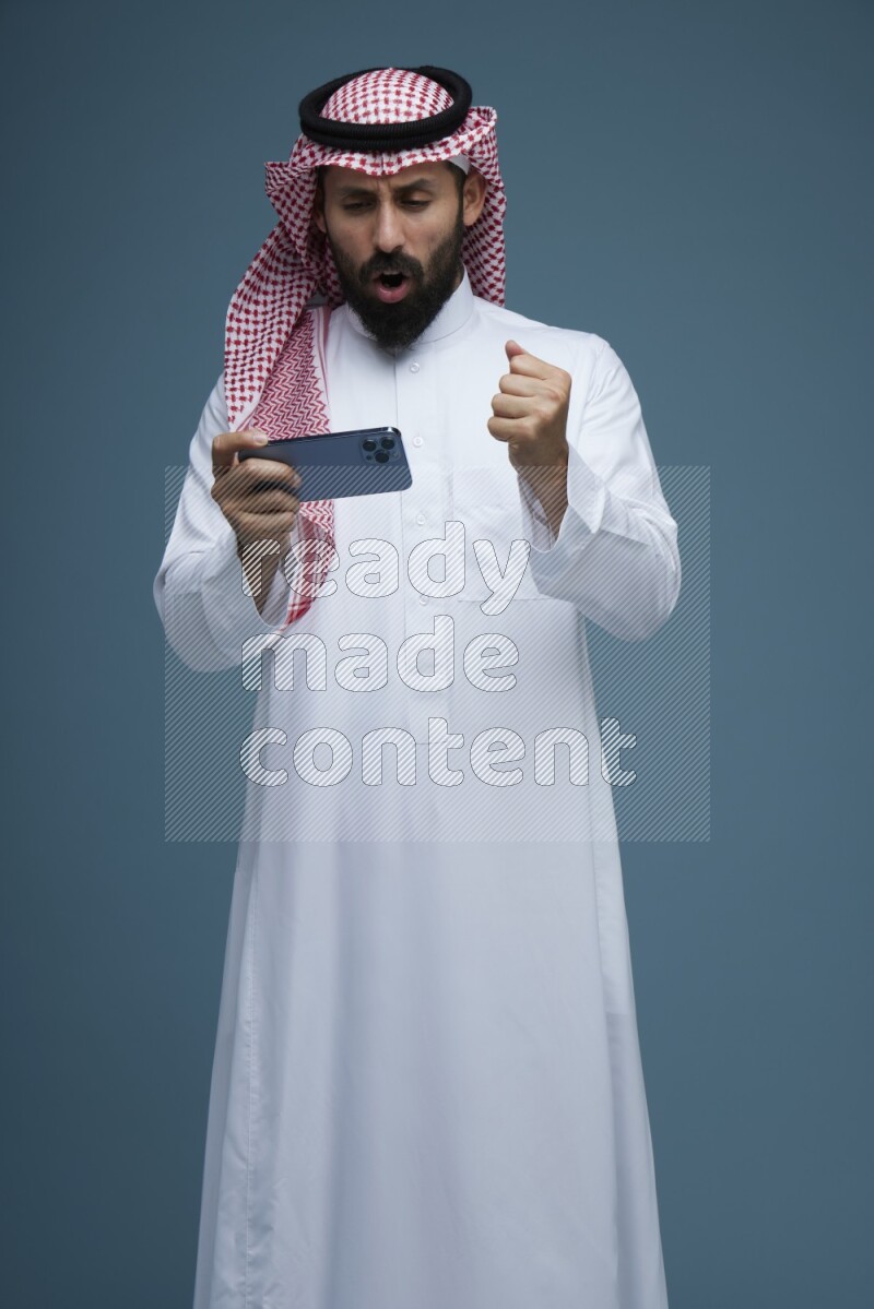 A man Playing a Game on his phone in a blue background wearing Saudi Thob with Shomag