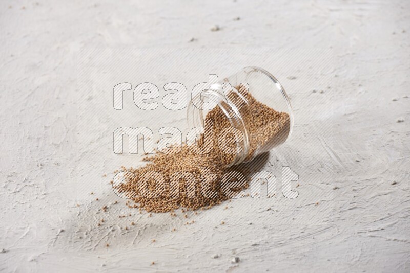 A glass jar full of mustard seeds and jar is flipped with seeds spread out on a textured white flooring