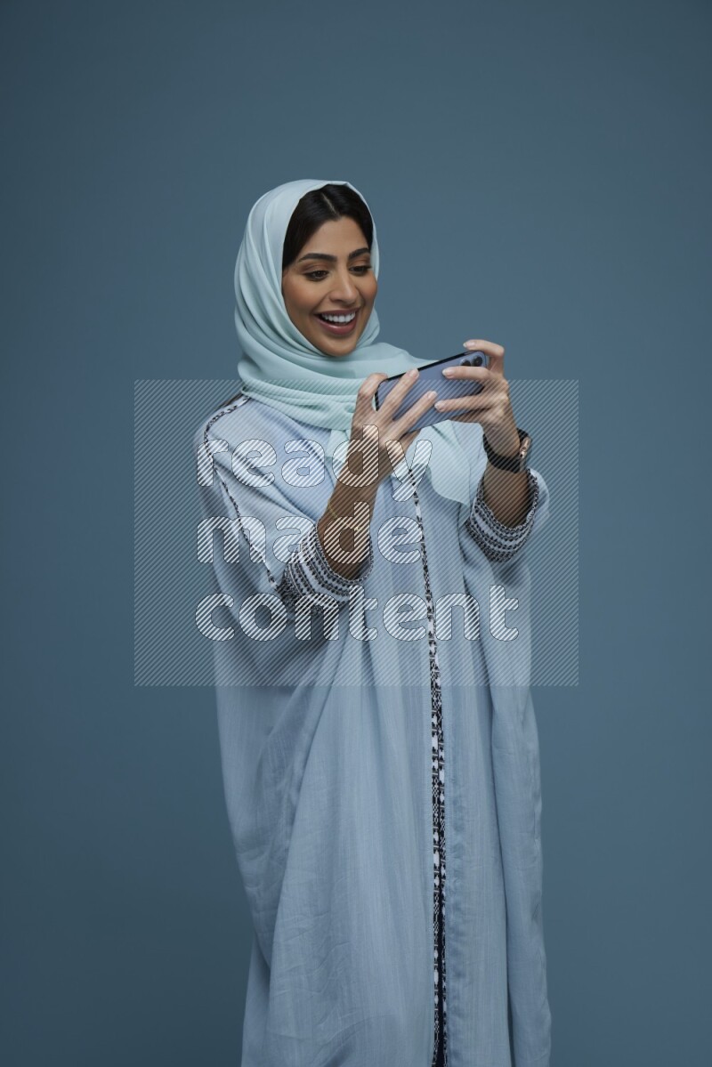 A Saudi woman Playing a Game on her phone on a blue background wearing a blue Abaya with hijab