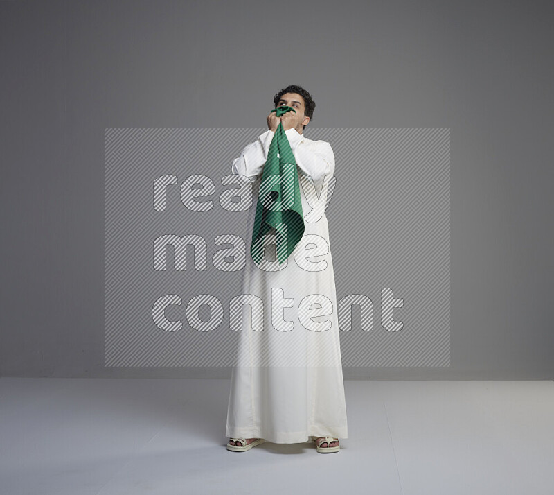 A Saudi man standing wearing thob kissing Saudi flag on gray background