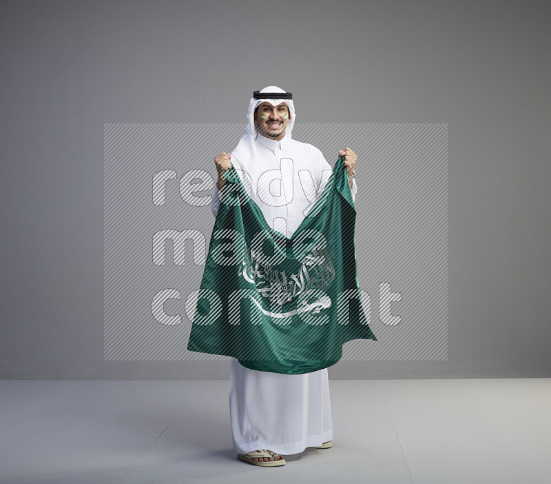 A Saudi man standing wearing thob and white shomag with face painting holding big Saudi flag on gray background