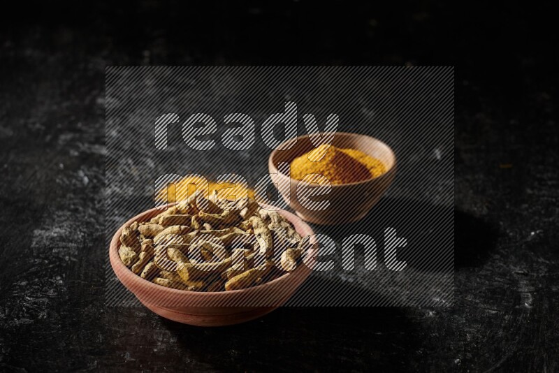 2 wooden bowls, one full of turmeric powder and the other full of dried turmeric whole fingers on a textured black flooring