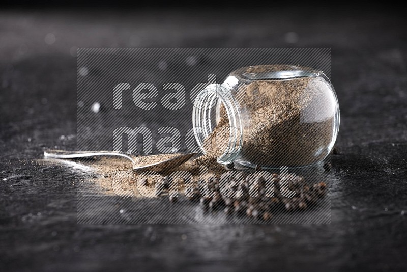 A flipped glass spice jar full of black pepper powder with a metal spoon full of powder and pepper beads on textured black flooring