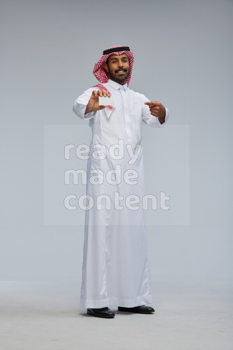 Saudi man wearing thob and shomag standing holding ATM card on gray background