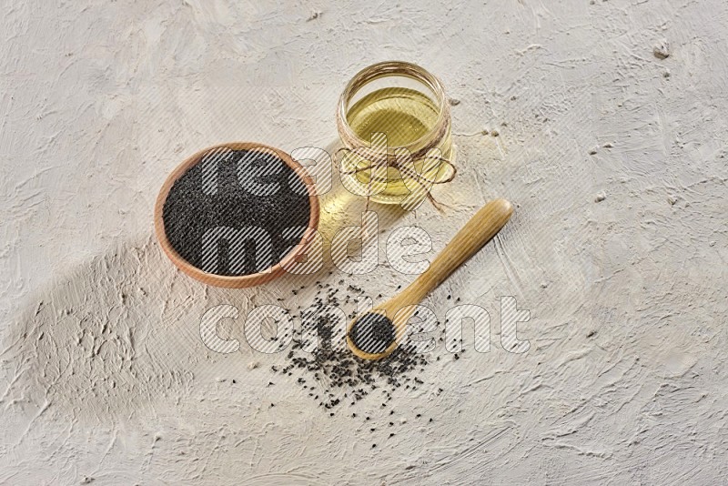 A wooden bowl and spoon full of black seeds and a glass jar of black seeds oil on a textured white flooring in different angles